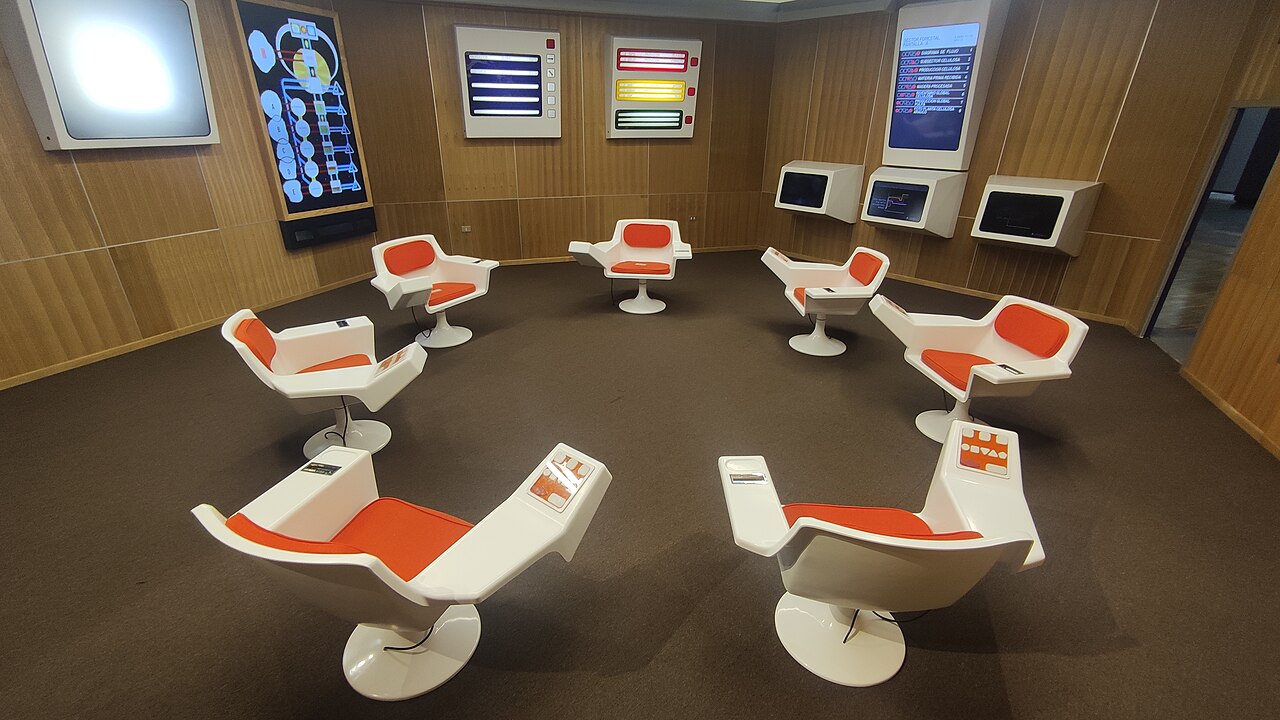 A circle of white swivel chairs with orange seats facing wall screens, a recreation of the Cybersyn operations room.