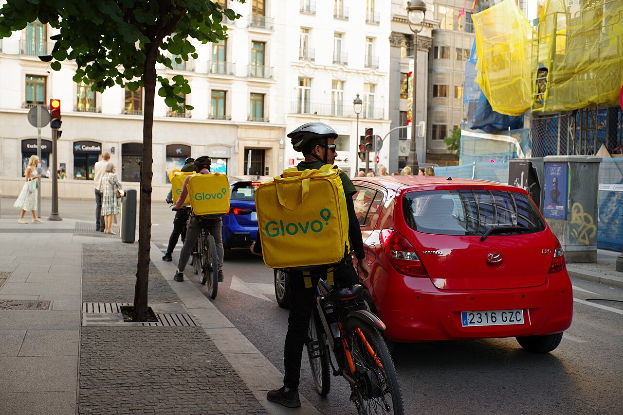 Glovo delivery riders with yellow backpacks waiting on bicycles in a Madrid street.