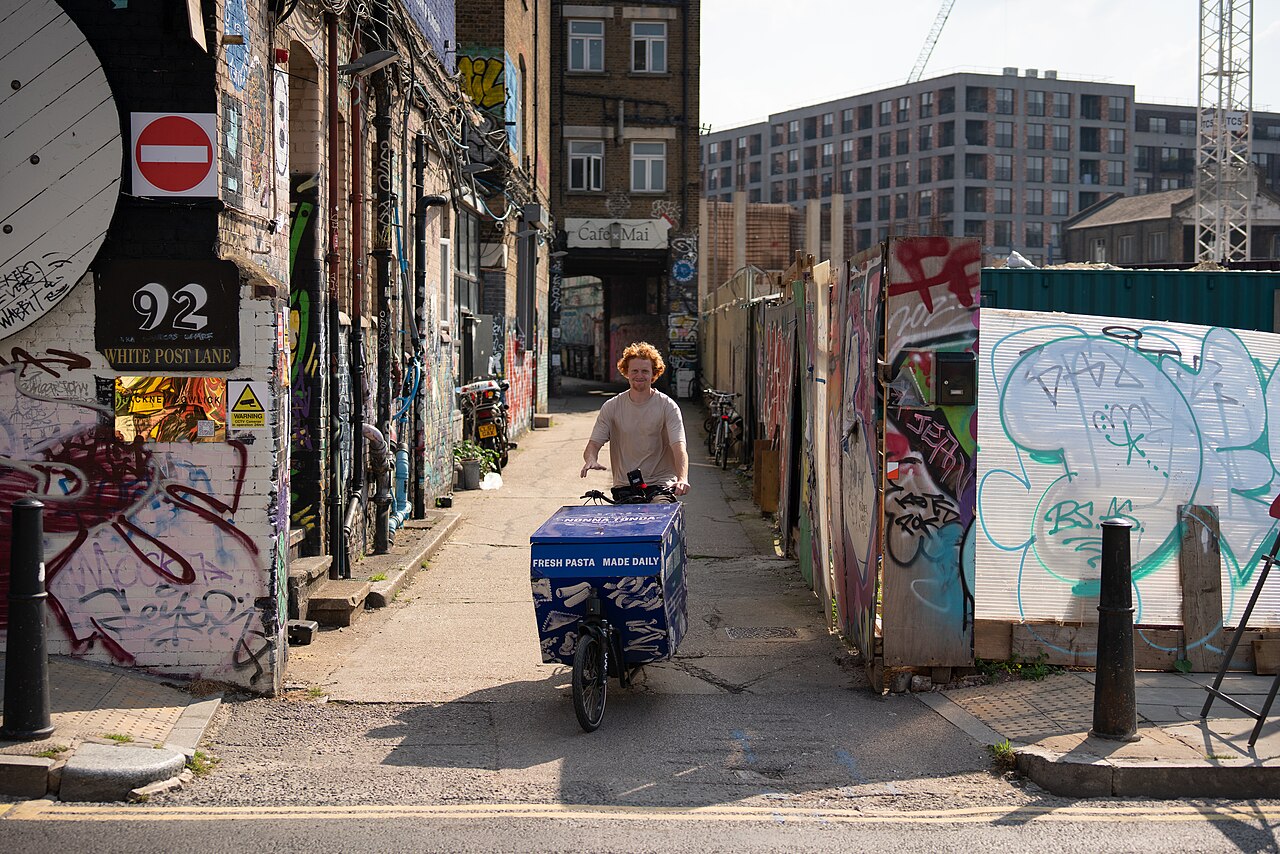 A rider on a loaded delivery cargo bike in a graffitied street in Hackney Wick, London.
