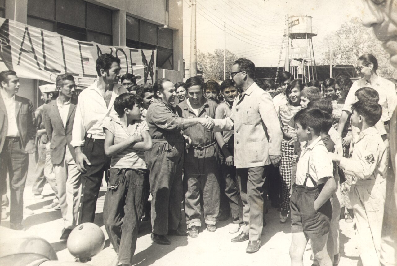 President Salvador Allende shaking hands with workers in overalls, among a crowd, in 1972.